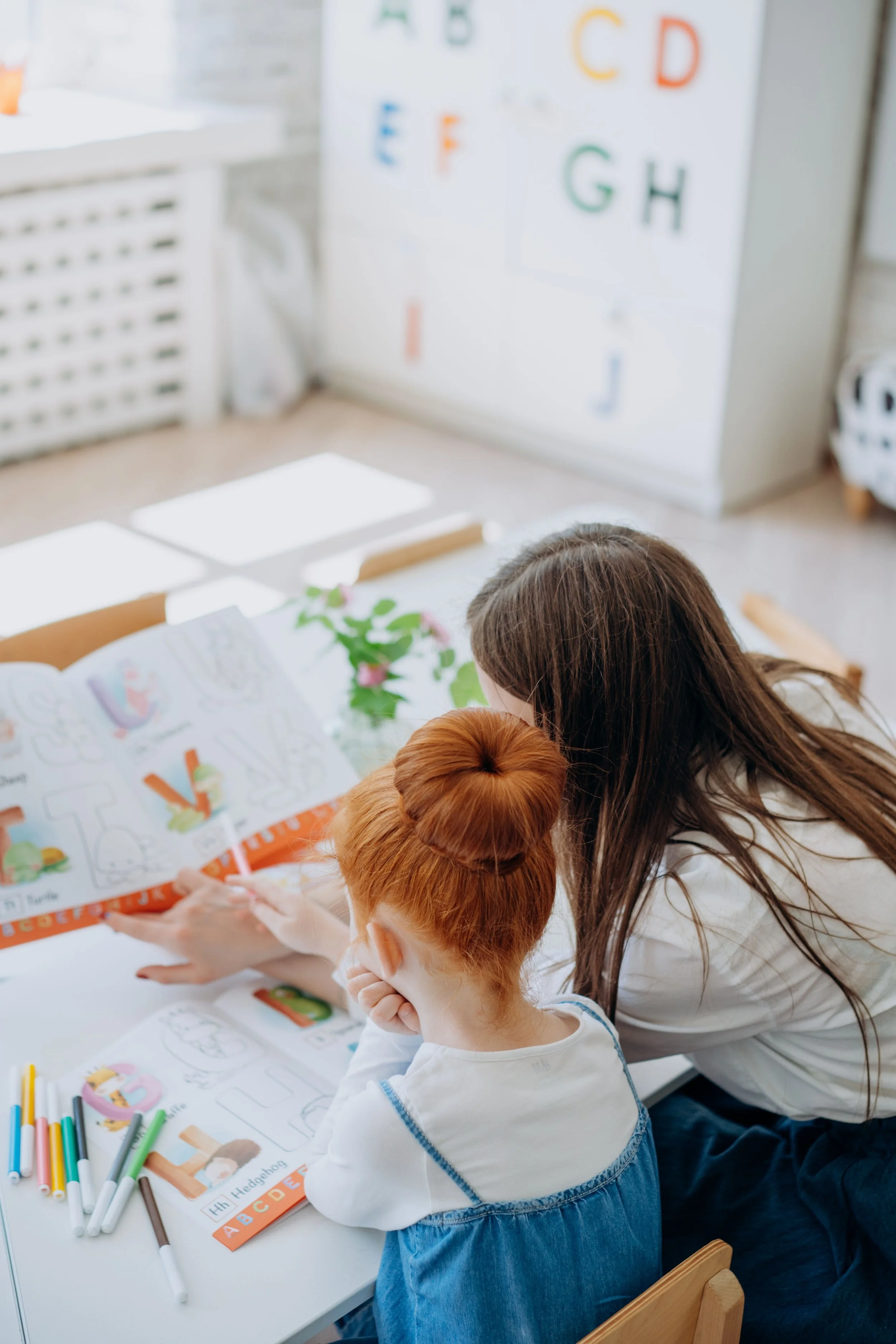 A woman and two children sitting at a table looking at coloring books with alphabet and animal illustrations, with colored markers on the table, in a bright room with a white cabinet with letters on the wall.