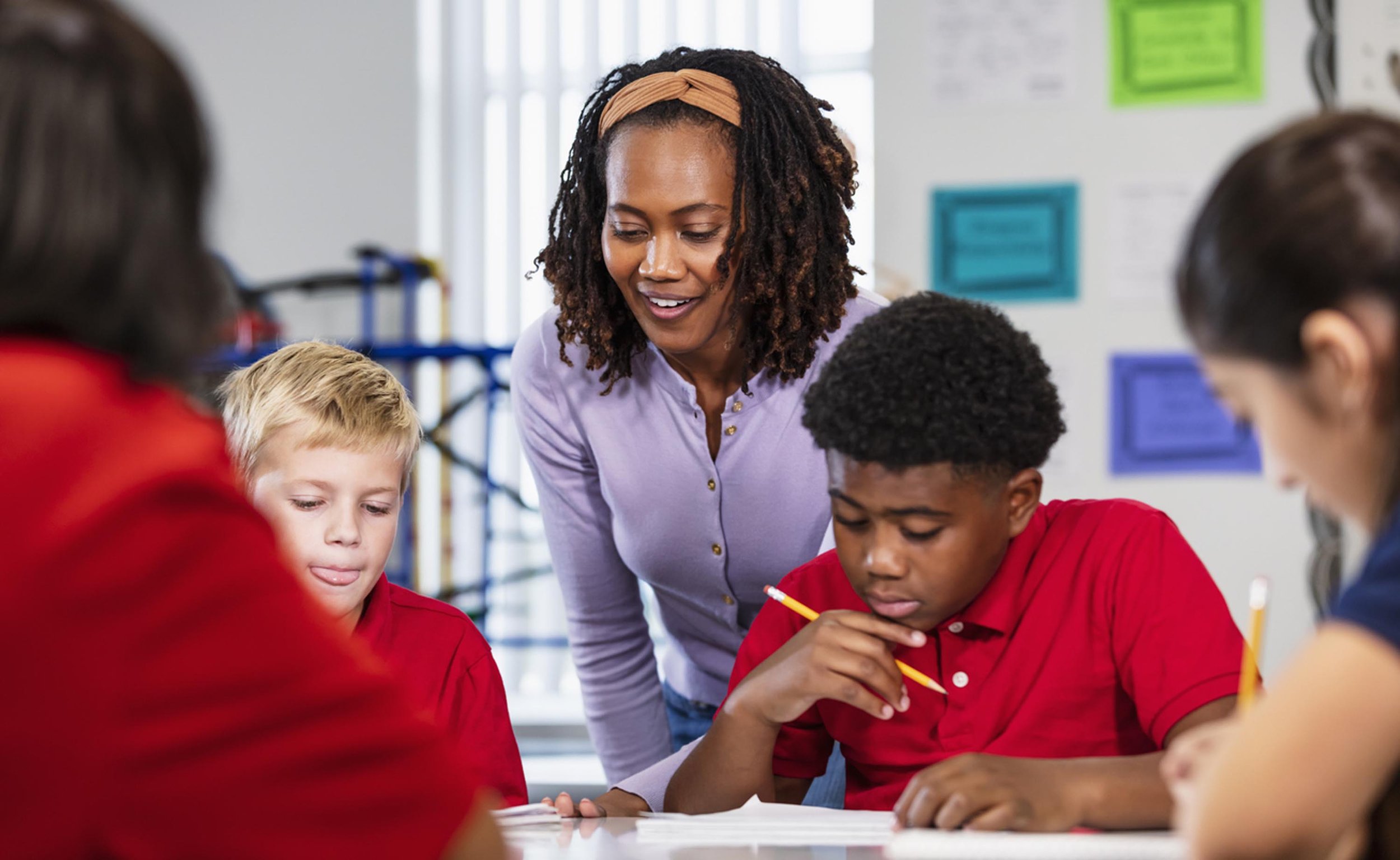 A teacher or instructor helping a group of students with a classroom activity. The students are focused, some with pencils, and wearing red shirts. The room has colorful decor on the walls.