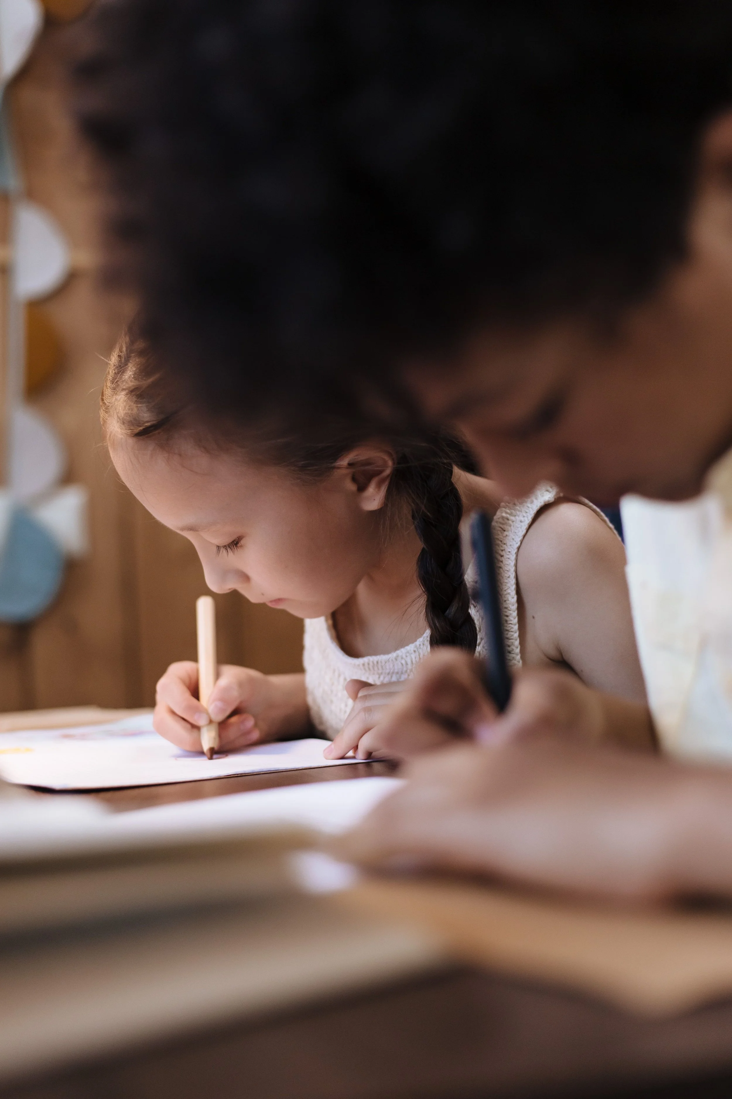A young girl with braided hair writing with a pencil at a table, with another person (possibly an adult) sitting next to her, both focused on their work.