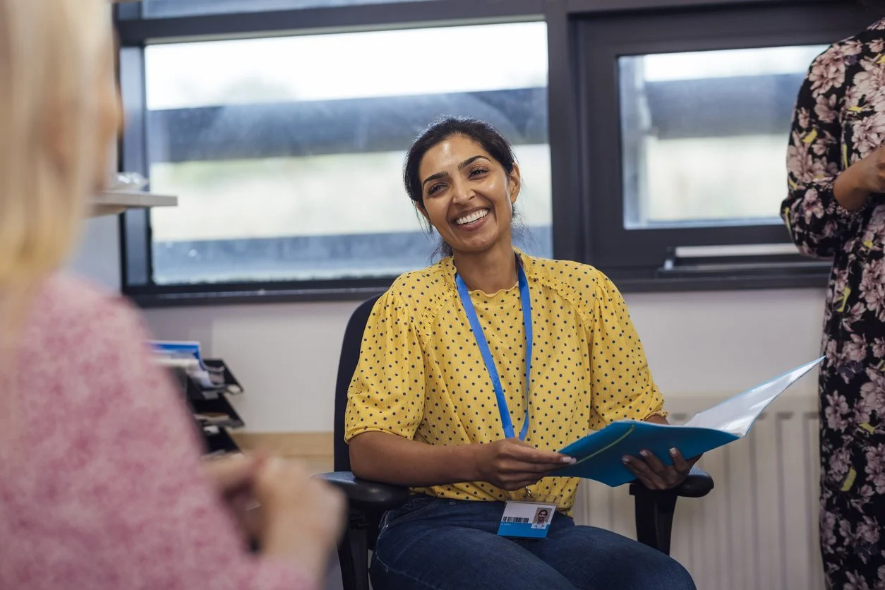 Woman with dark hair in a bun, wearing a yellow polka-dotted top, smiling and holding blue folders, sitting in an office with large windows, engaging with two other women.