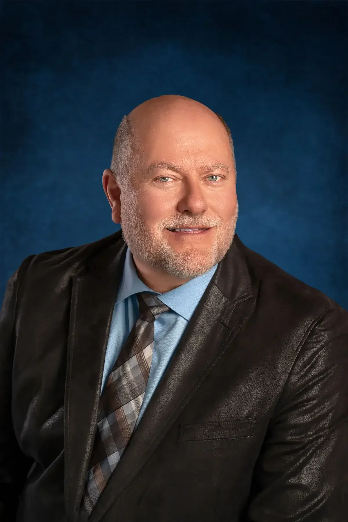 Portrait of a middle-aged man with light skin, blue eyes, and a beard, wearing a black leather jacket, a light blue shirt, and a plaid tie, against a dark blue background.