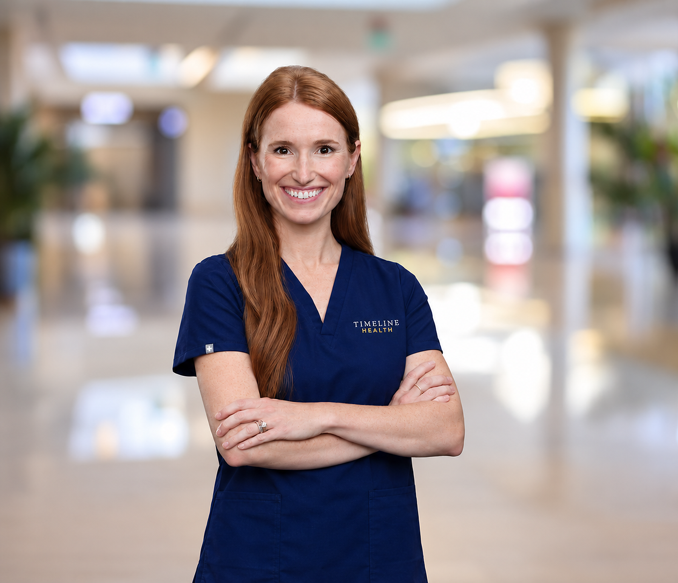 Founder, Charlotte MacDonald, wearing navy Timeline Health medical scrubs, standing with arms crossed in a bright indoor setting.
