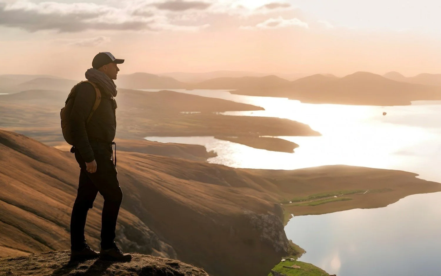 A man standing on a mountain ridge overlooking a scenic landscape with lakes, rolling hills, and distant mountains.