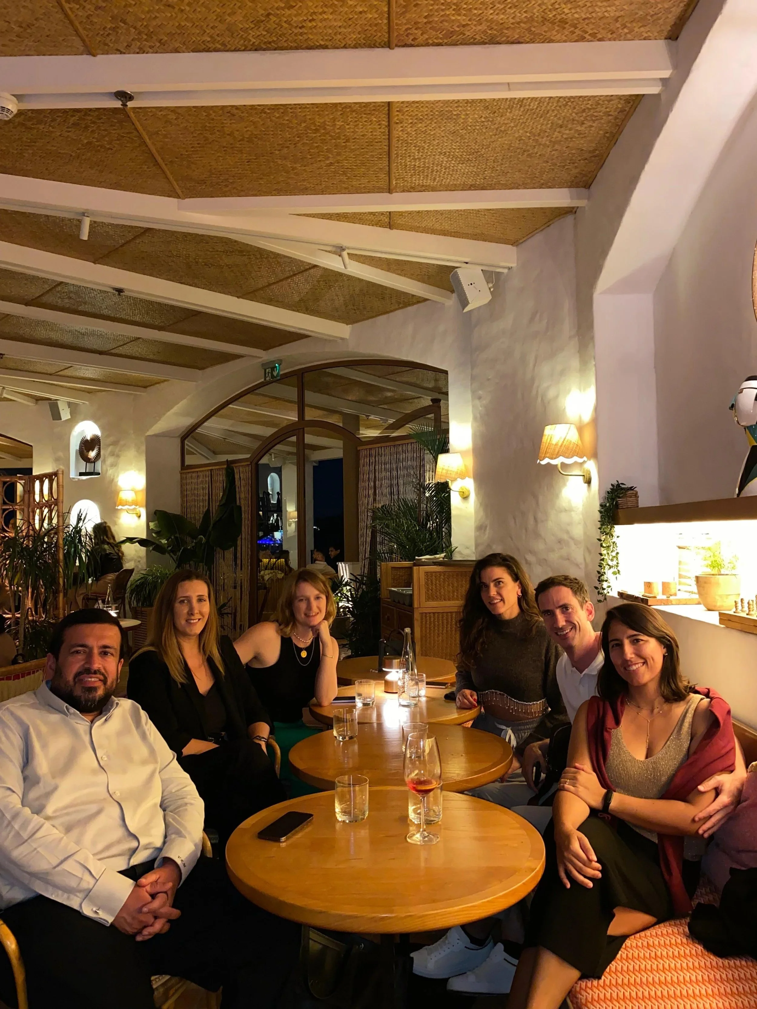 Group of seven people, four women and three men, sitting around a wooden table in a restaurant or cafe, smiling and posing for the photo. The setting has warm lighting, plants, and decorative wall lamps.
