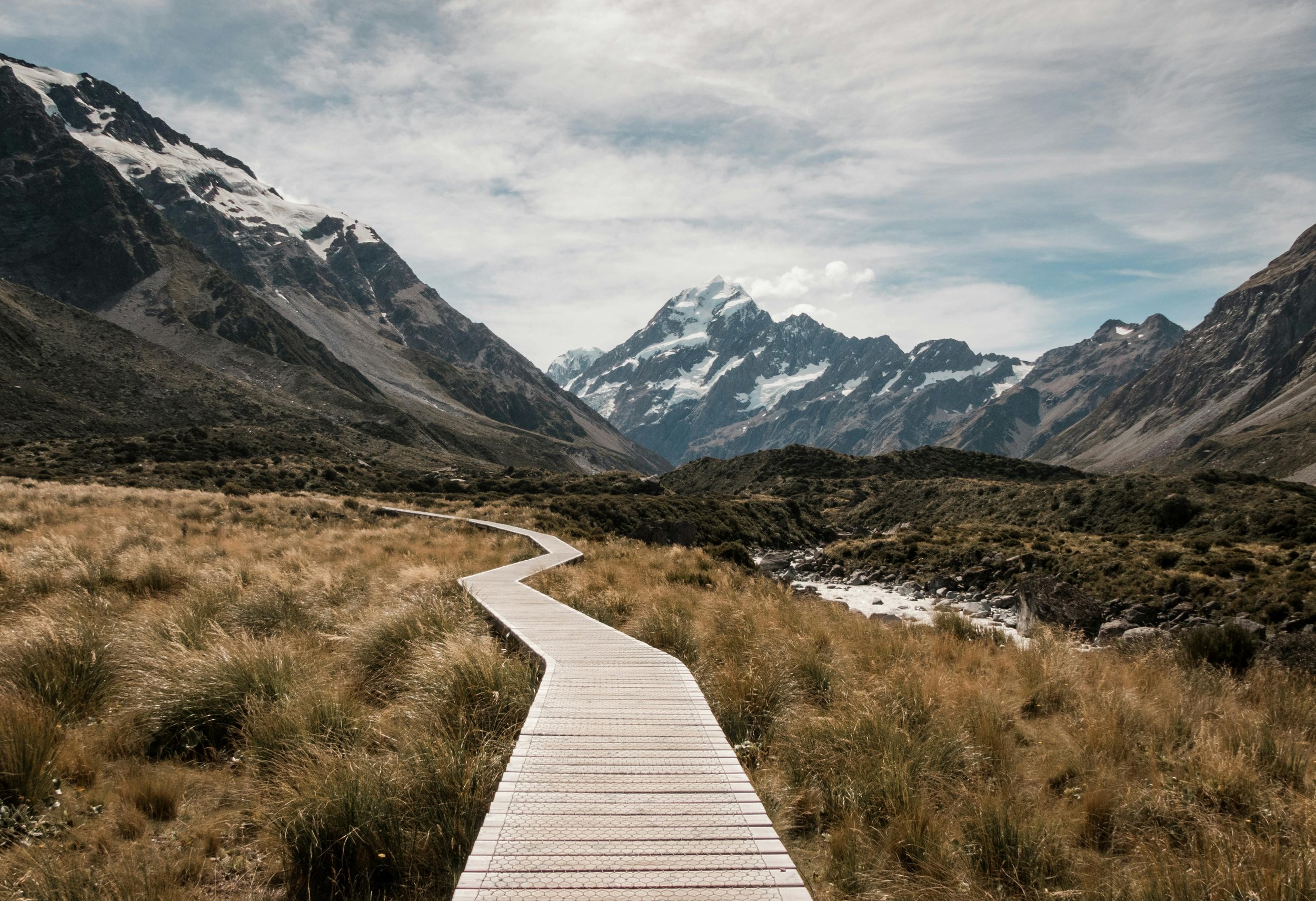 A wooden walkway through grassy terrain leads towards snow-capped mountains under a cloudy sky.