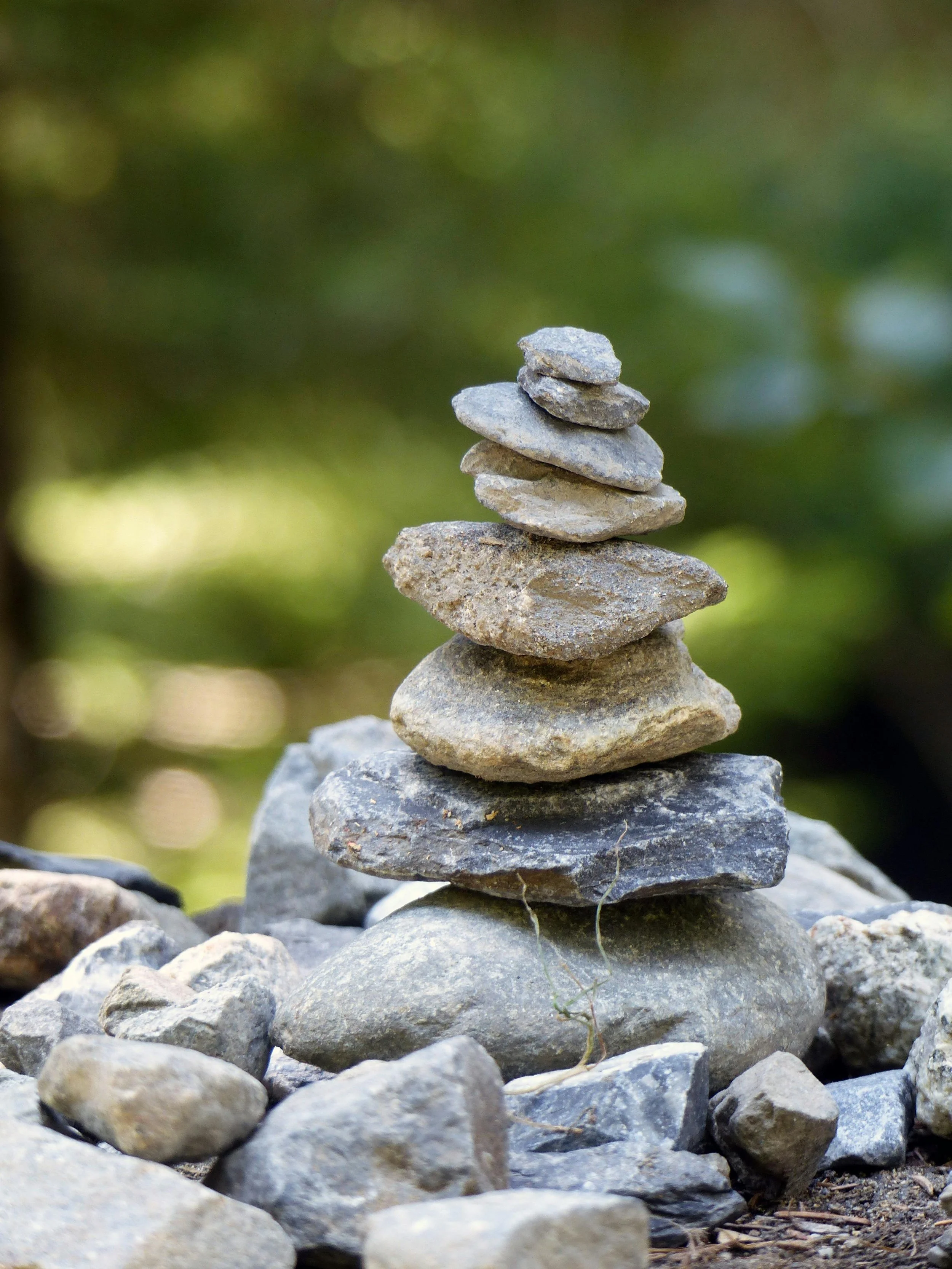 A stack of seven smooth, flat stones balanced on top of each other on a rocky ground with a blurred green background.