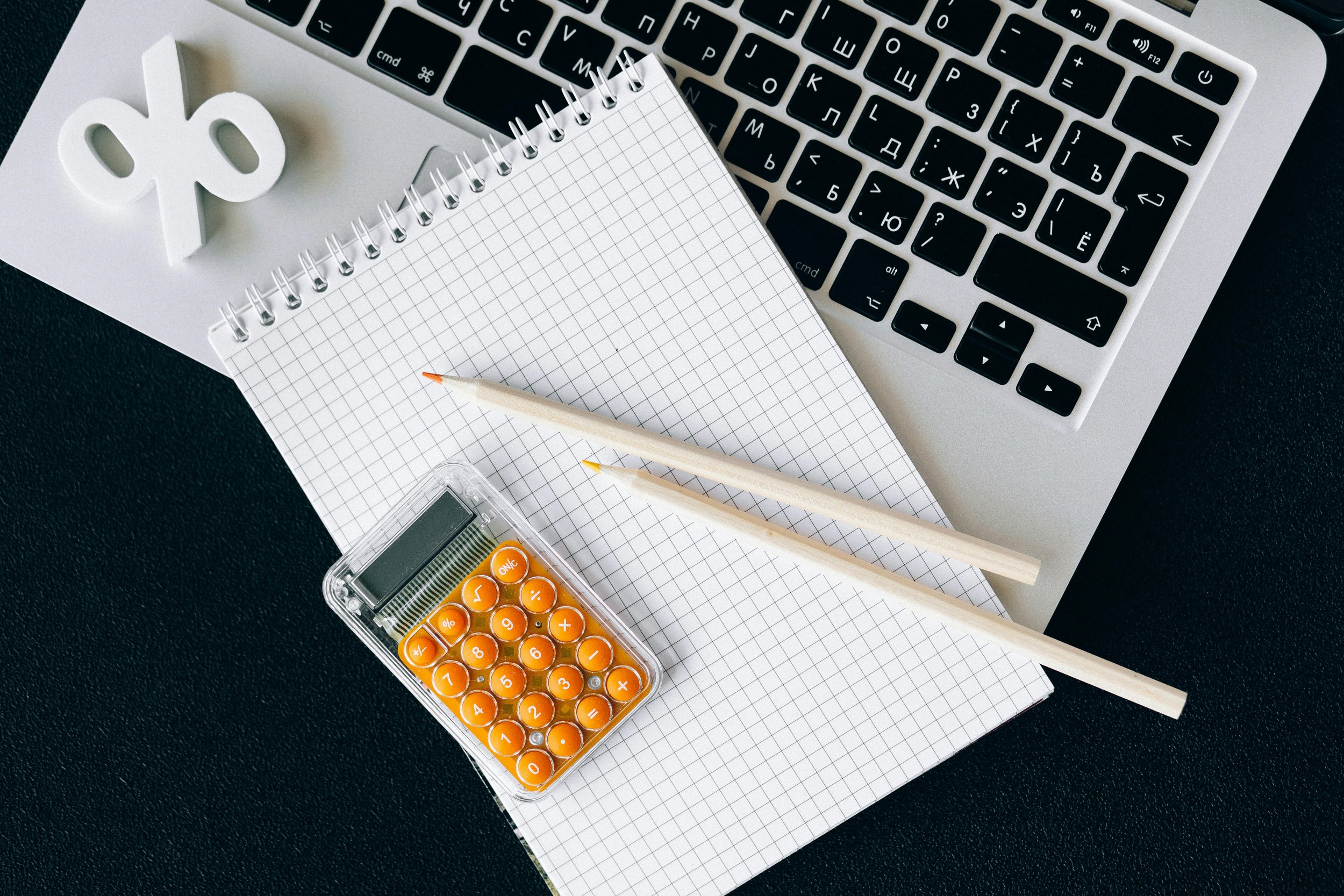A workspace with a laptop keyboard, a small spiral notepad, a sheet of graph paper, two pencils, and a container of orange and yellow calculator batteries.