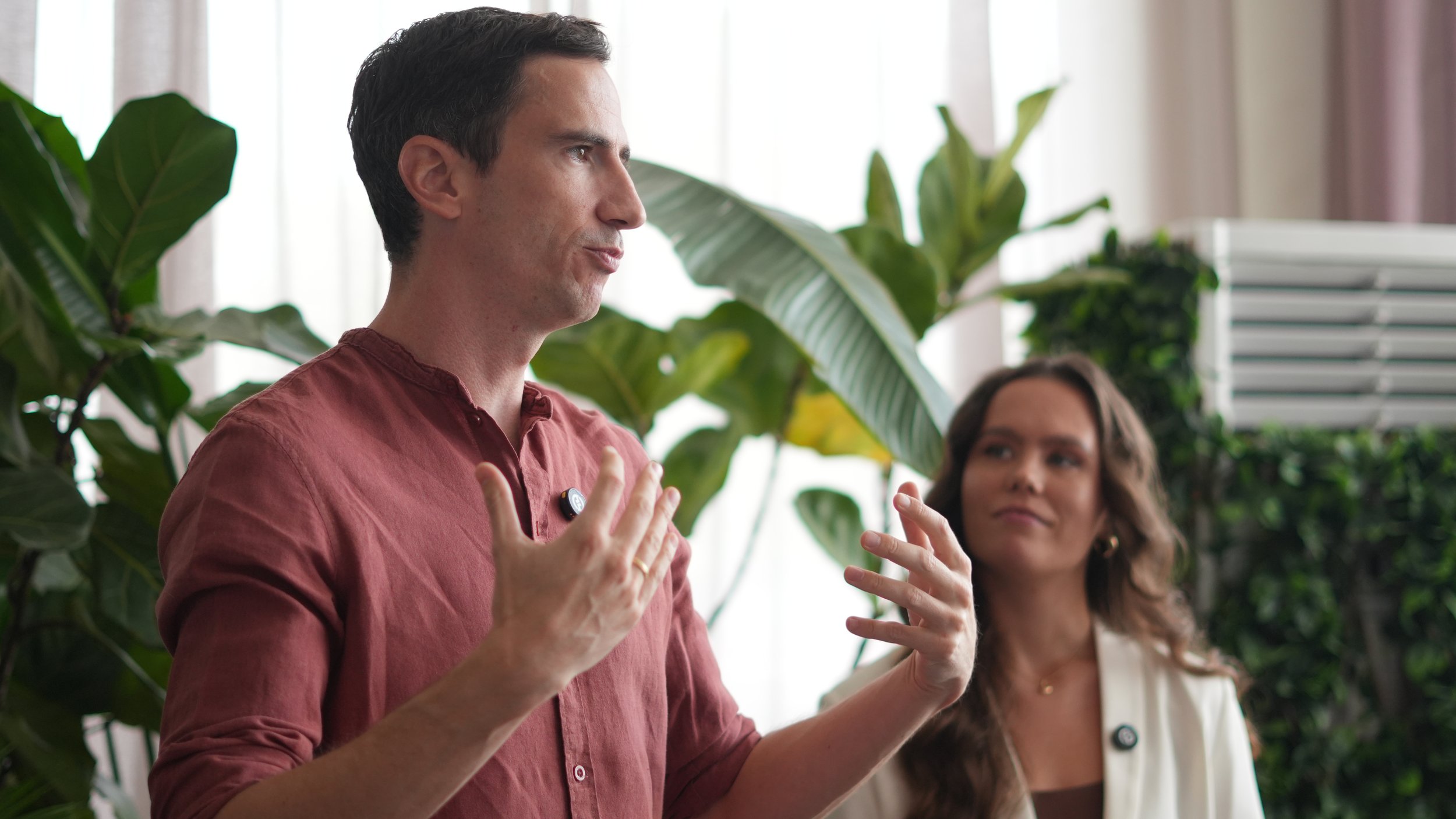 A man with dark hair and a pink shirt speaking with hand gestures, standing beside a woman with curly hair and a white blazer, in front of green plants and natural light.