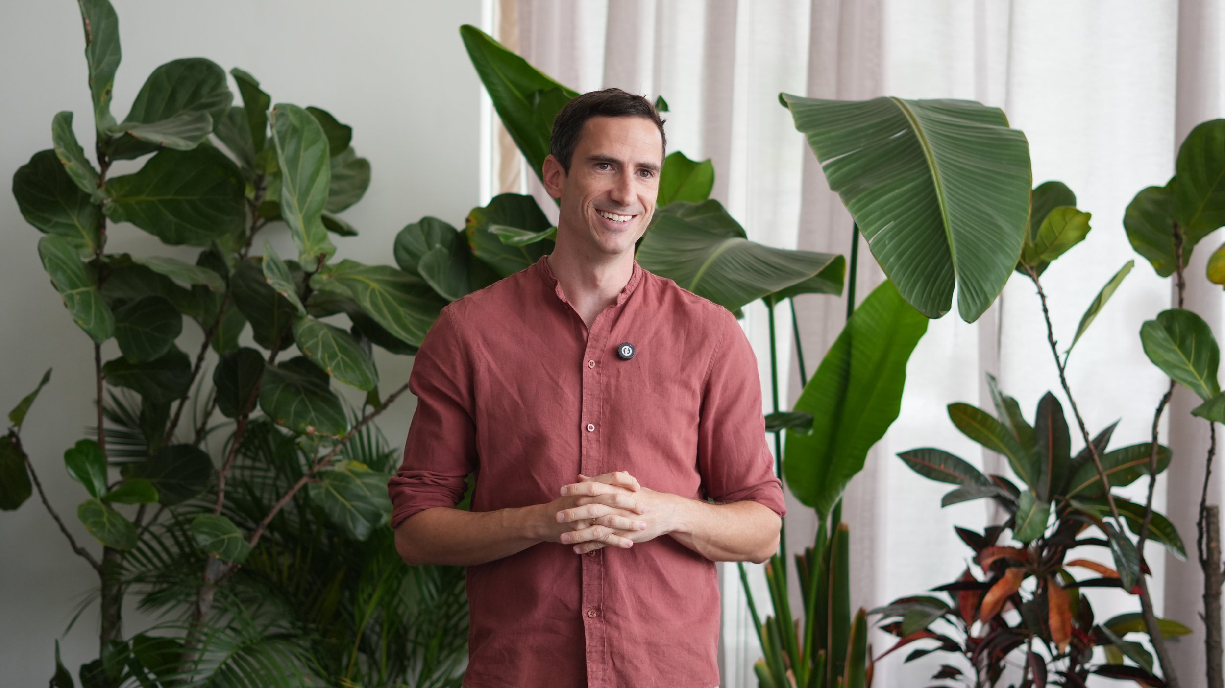 A smiling man in a salmon-colored shirt standing in front of large green houseplants with sheer white curtains in the background.
