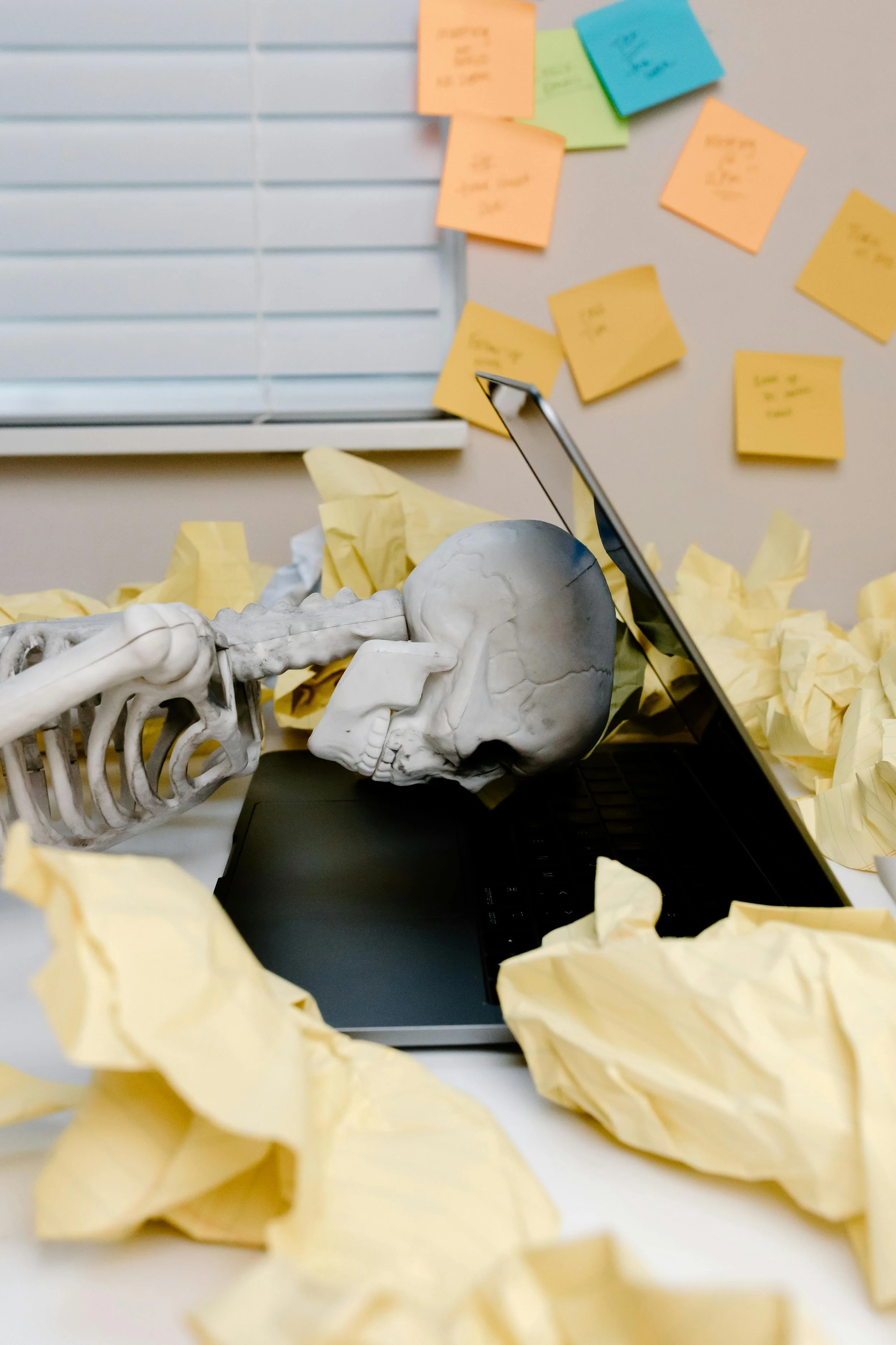 A human skeleton model lying on a desk surrounded by crumpled yellow sticky notes and a tablet. In the background, there are colorful sticky notes on a wall, some with handwritten notes.