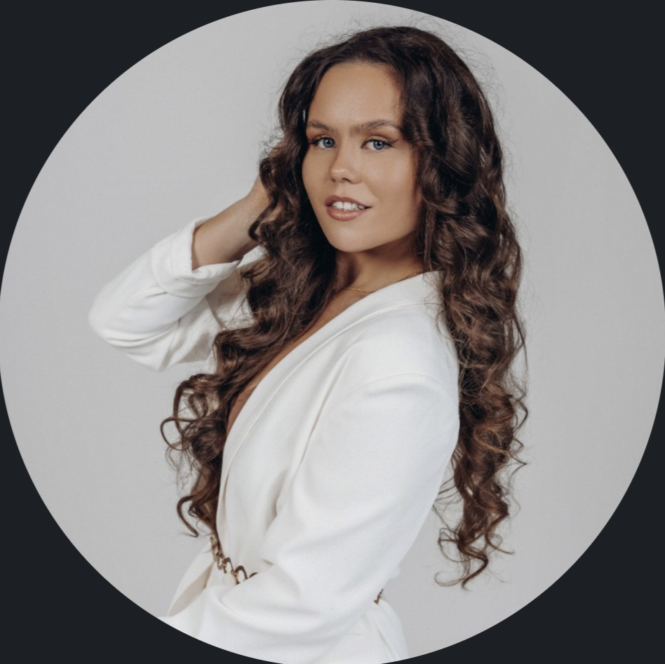 A woman with long, curly brown hair wearing a white blazer, posing with one hand near her head against a gray background.
