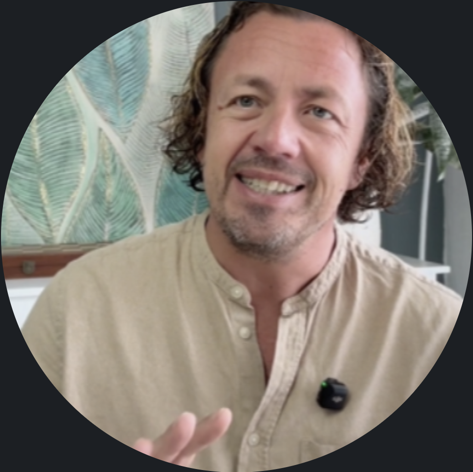 A man with curly brown hair and a beard smiling and talking, wearing a beige button-up shirt with a microphone clipped to it, in front of a leafy patterned background.