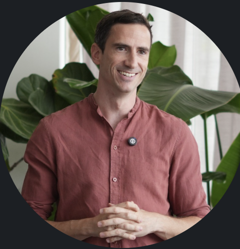 A man with short dark hair smiles while sitting in front of large green houseplants.
