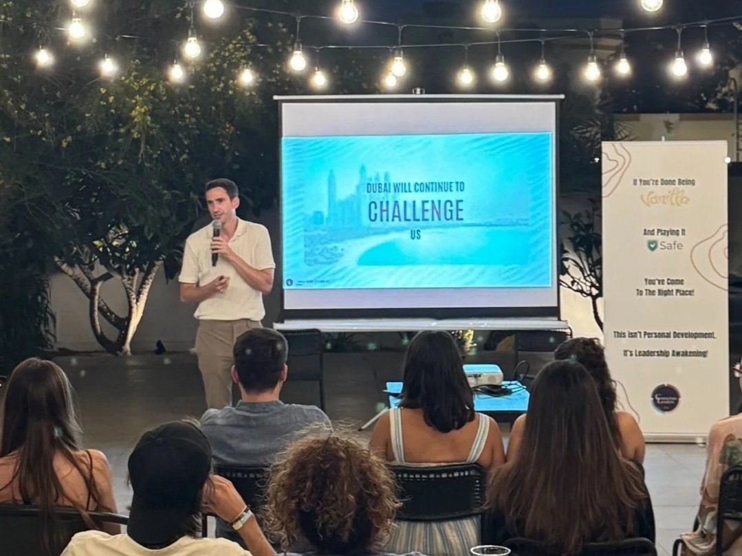 A man giving a presentation in front of a slide that says 'Dubai will continue to challenge us.' The audience is seated facing the presenter, with string lights hanging above and trees in the background.