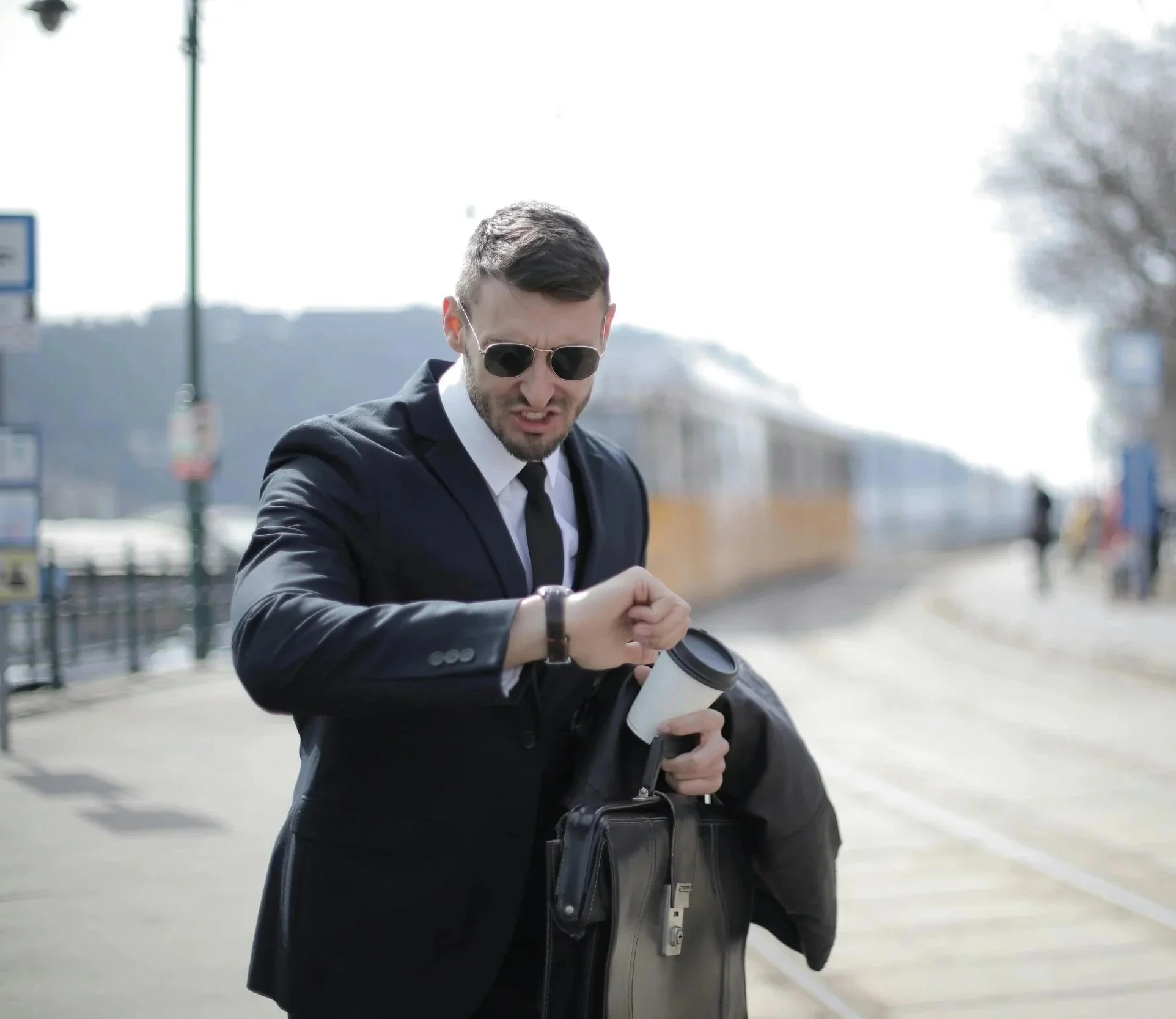 A man in a suit and sunglasses looking at his watch outside on a sunny day, holding a coffee cup, with a blurred urban background.