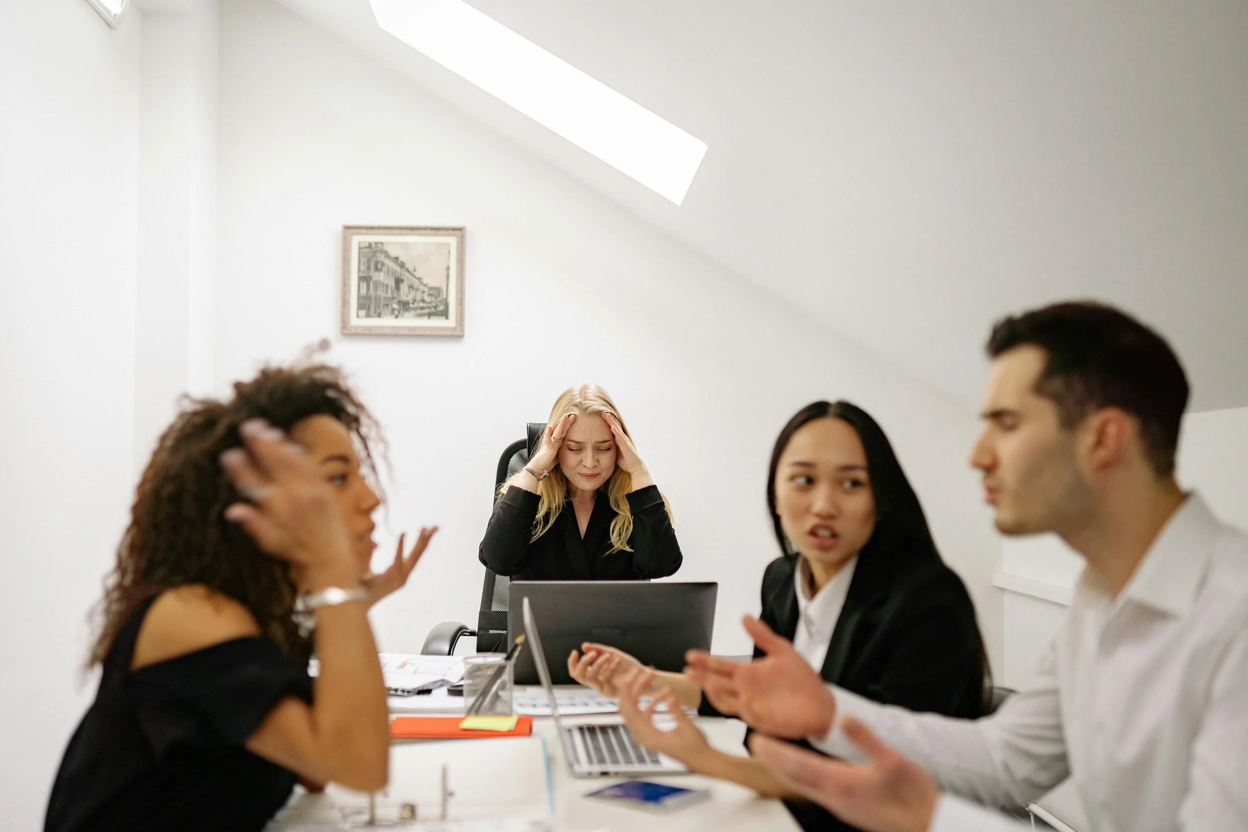 A group of people in a meeting or discussion, with a woman in the center holding her head and looking distressed, while others are engaged in conversations."