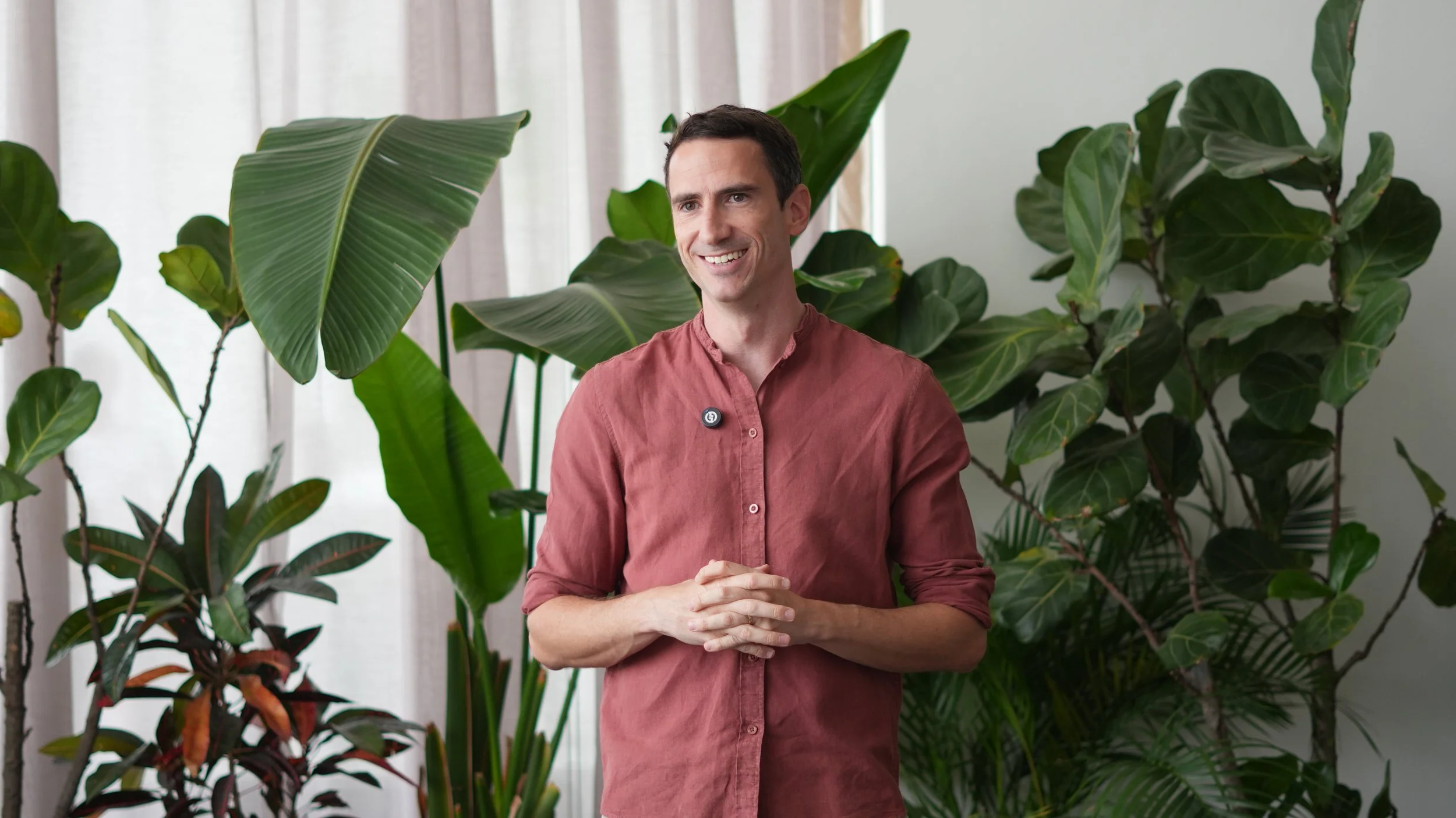 A man with dark hair wearing a reddish-brown button-up shirt stands among large green indoor plants, smiling.