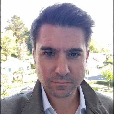A man with dark hair and a serious expression takes a selfie outdoors with trees and houses in the background.