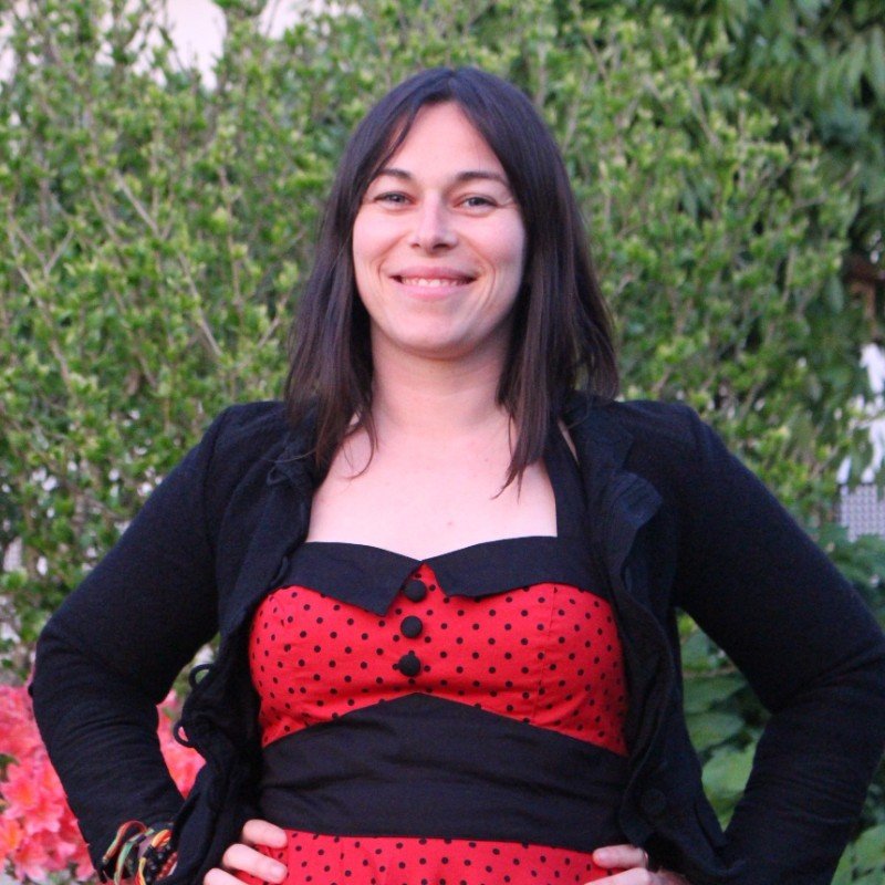 A woman smiling outdoors with green bushes in the background, wearing a red and black polka dot dress and a black jacket.