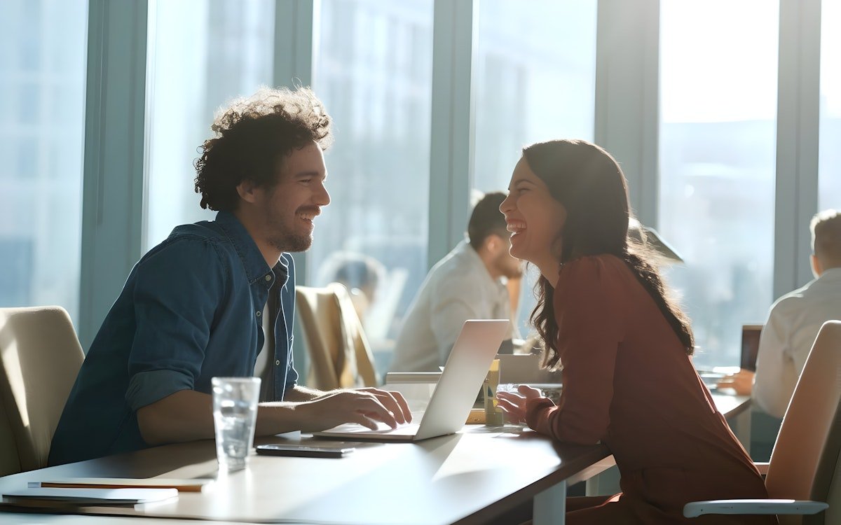 Two people sitting at a table in a modern office, smiling and laughing together.
