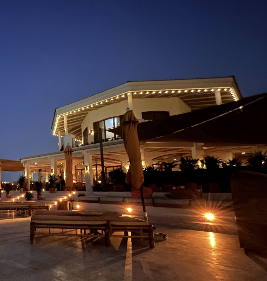 A large, multi-story house or restaurant with white exterior walls and string lights along the roofline, illuminated at night with outdoor seating and umbrellas on a patio.