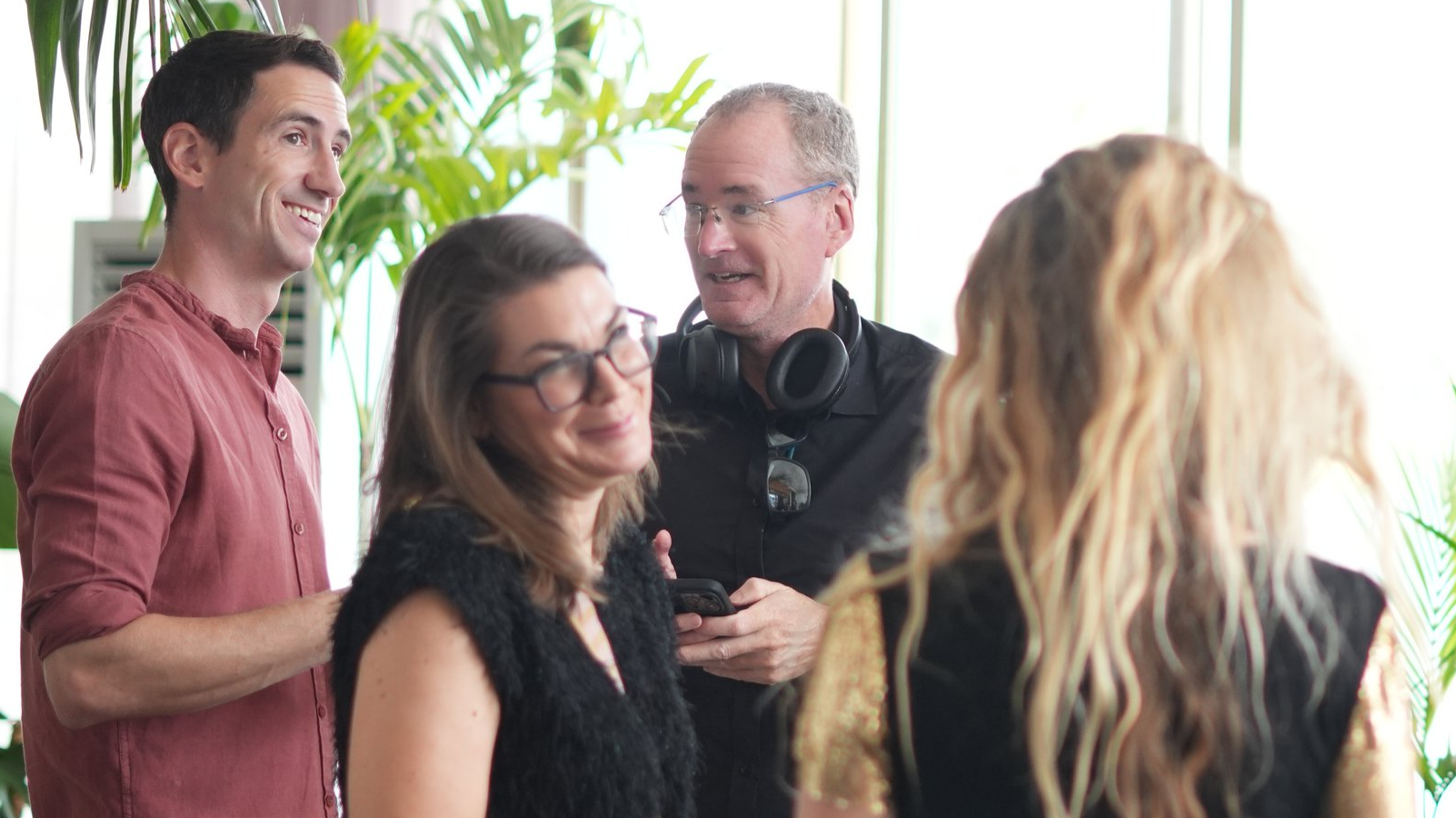 Four people in a bright, indoor space chatting and smiling, with plants and large windows in the background.