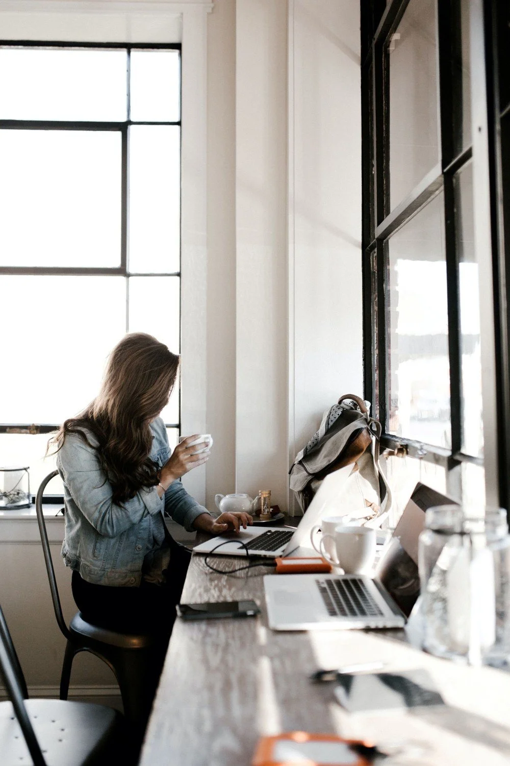 A woman with long brown hair sitting at a cluttered wooden desk in a room with large windows, using a laptop and holding a cup.