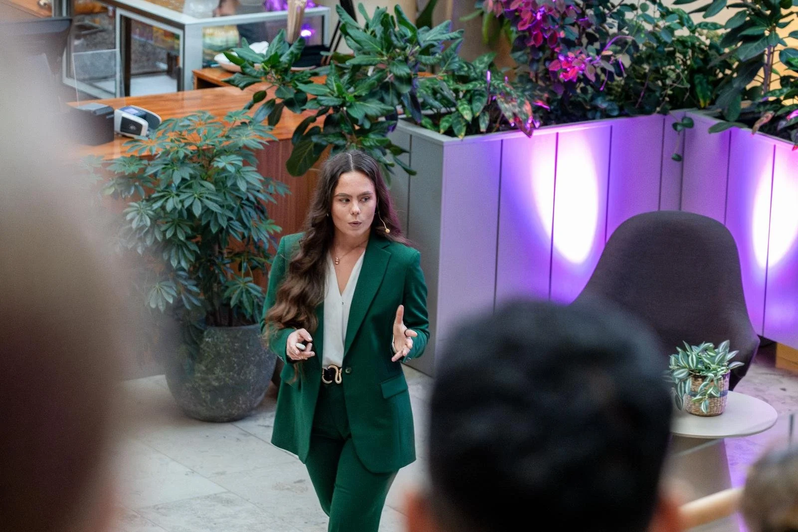A woman in a green suit giving a presentation or speech in a modern indoor space with plants and purple lighting.