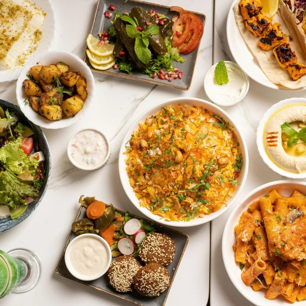 An assortment of Middle Eastern dishes on a white marble table, including rice pilaf, hummus, salad, grilled meat skewers, falafel with tahini, and various vegetables and dips.