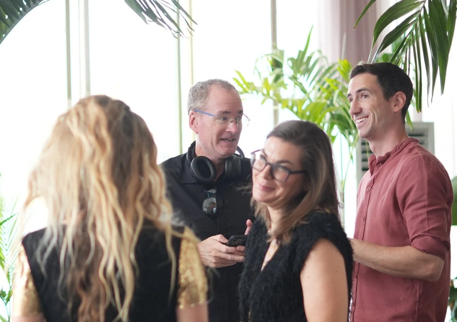 Group of four people talking and smiling indoors, with large green plants and natural light coming through windows in the background.