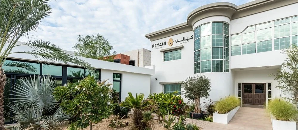 Modern white building with large glass windows and desert landscaping, sign reading 'Keyani' in English and Arabic.