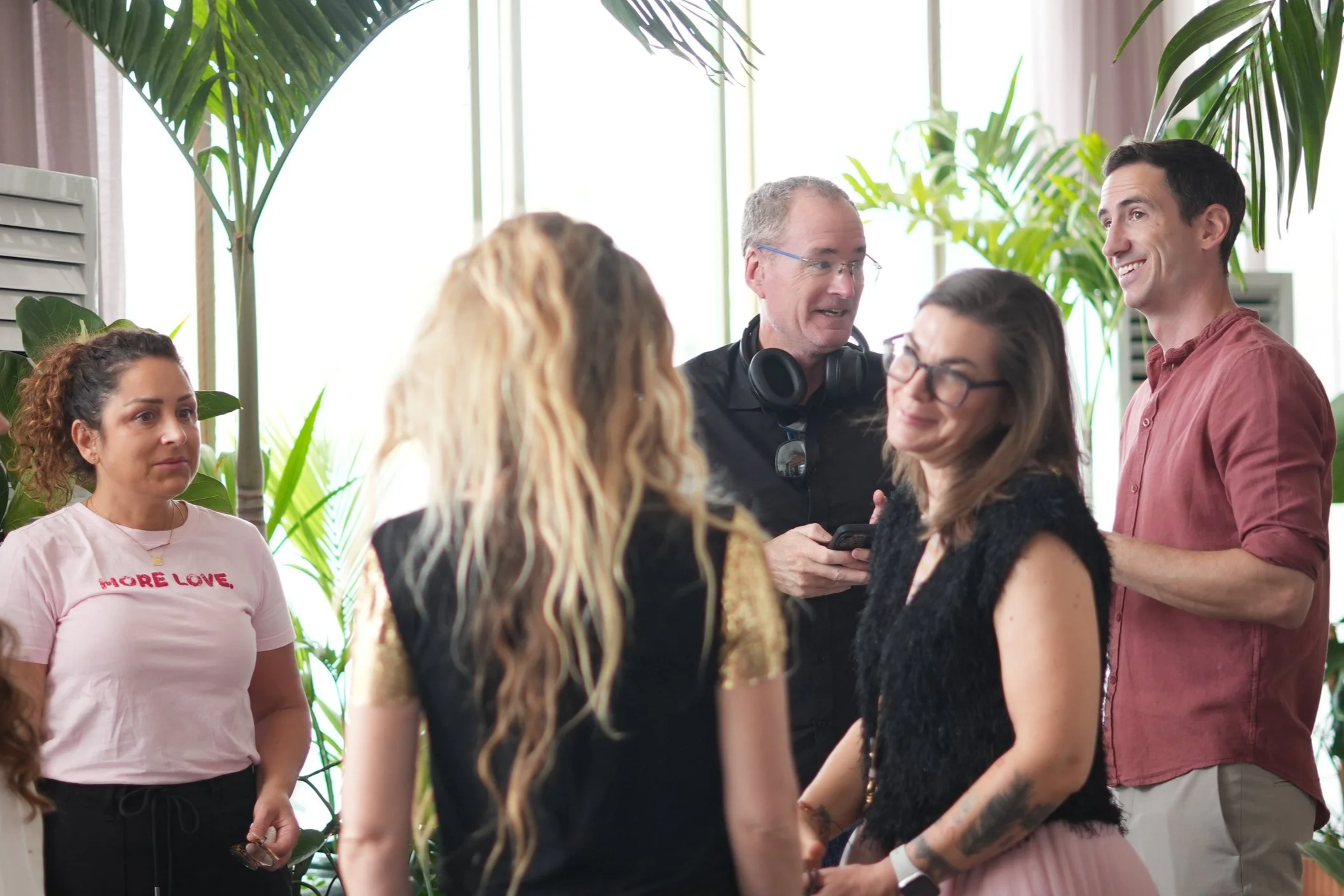 A group of diverse people are gathered indoors, engaging in conversation with a smiling man holding a cellphone, surrounded by green plants and natural light.