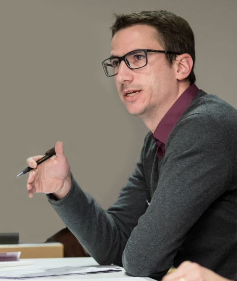 A man with glasses, dark hair, and a beard wearing a gray sweater over a maroon shirt, sitting at a desk and gesturing with a pen in his hand.