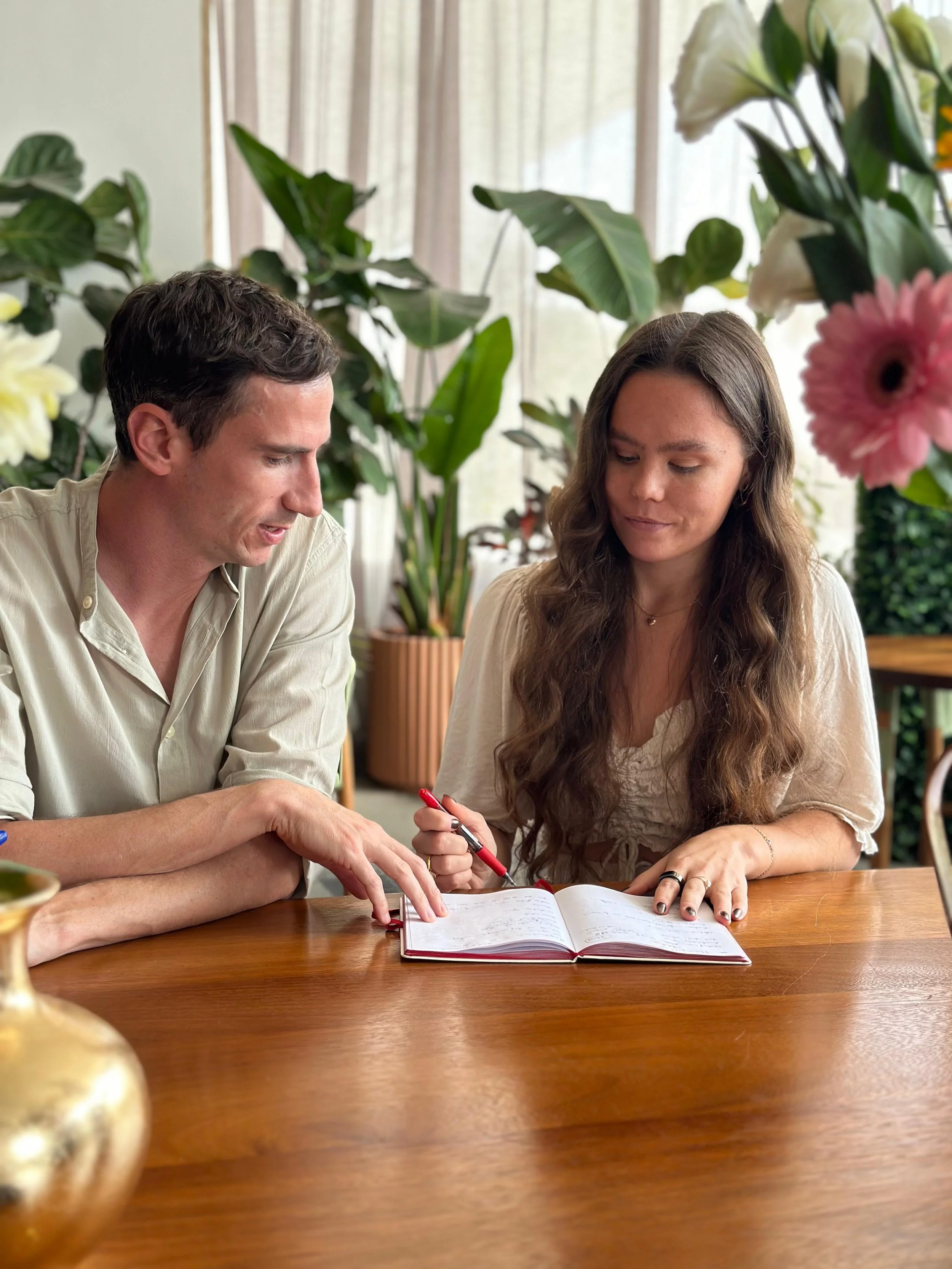 A man and woman sit at a wooden table, looking at an open notebook together. The man has dark hair and is wearing a light-colored shirt, pointing at the notebook. The woman has long, wavy brown hair, wearing a light top, and holding a red pen. They are in a room with large green plants and sheer curtains in the background.