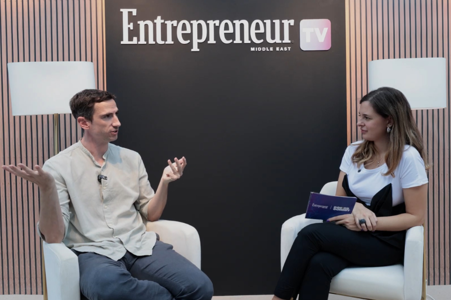 Two people sitting in chairs having an interview, backstage of Entrepreneur TV Middle East, with a black backdrop and branding. The man on the left is speaking with a gesture, and the woman on the right is listening, holding notes.