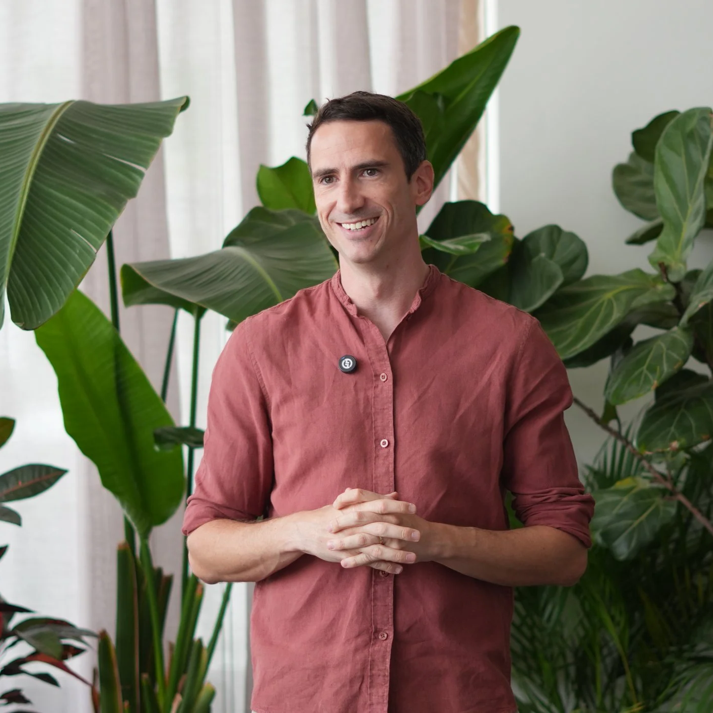 A smiling man with dark hair wearing a rust-colored button-up shirt standing in front of large green plants and light-colored curtains.