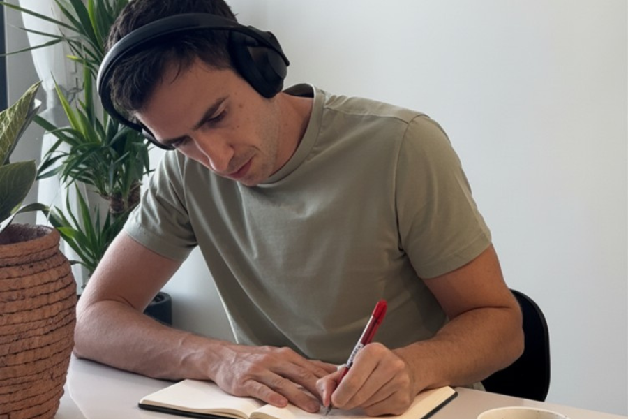 Young man sitting at a desk, wearing headphones, writing in a notebook with a red pen, next to potted plants.