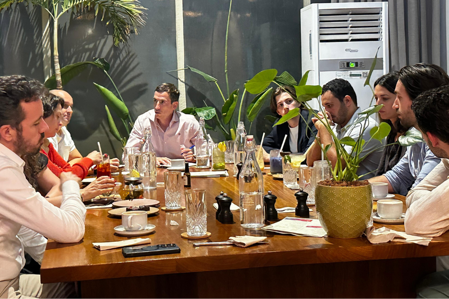A group of people sitting around a wooden table having a meeting in a modern restaurant or cafe with plants and an air conditioner in the background.