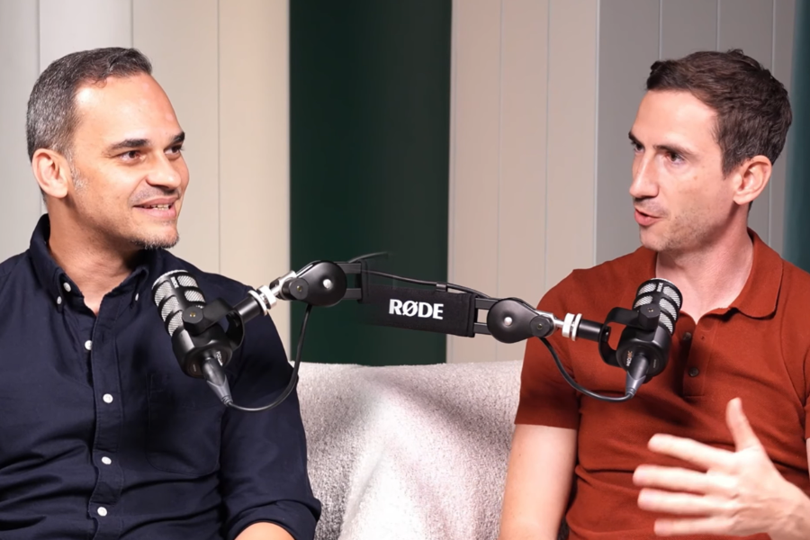 Two men sitting on a beige sofa having a conversation with microphones in front of them, podcast studio setting.