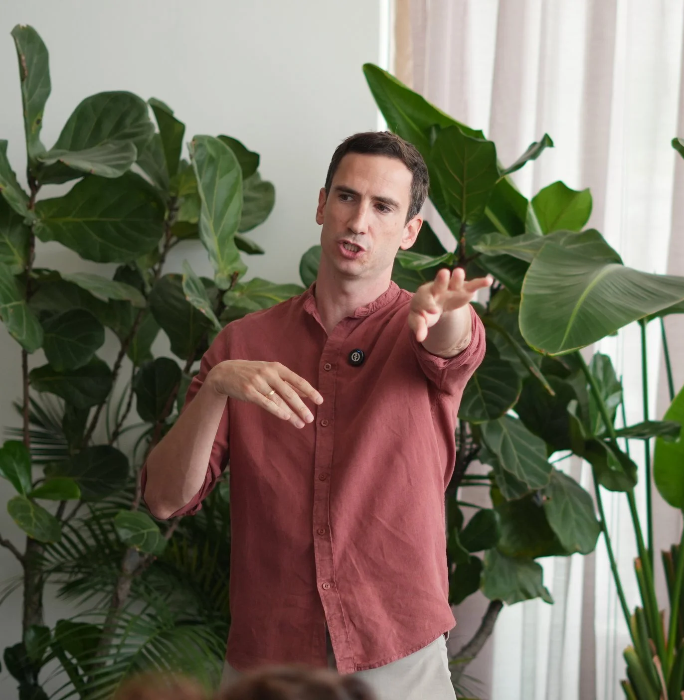 A man in a reddish-brown shirt speaking and gesturing with his hands in front of large green plants and a window with light-colored curtains.