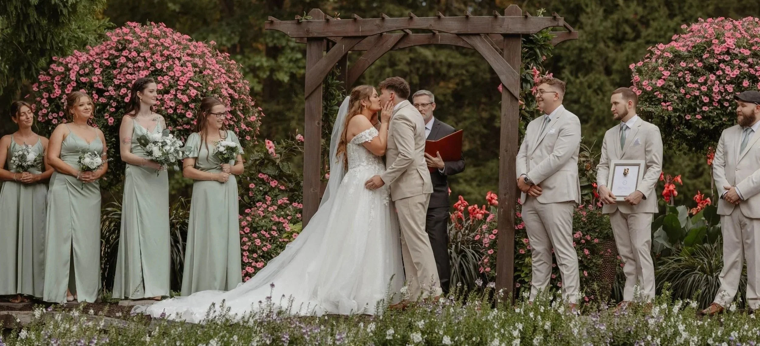 A wedding ceremony outdoors with a bride and groom kissing under an wooden arch, surrounded by bridesmaids and groomsmen, all dressed in formal attire, with pink flowers and greenery in the background.