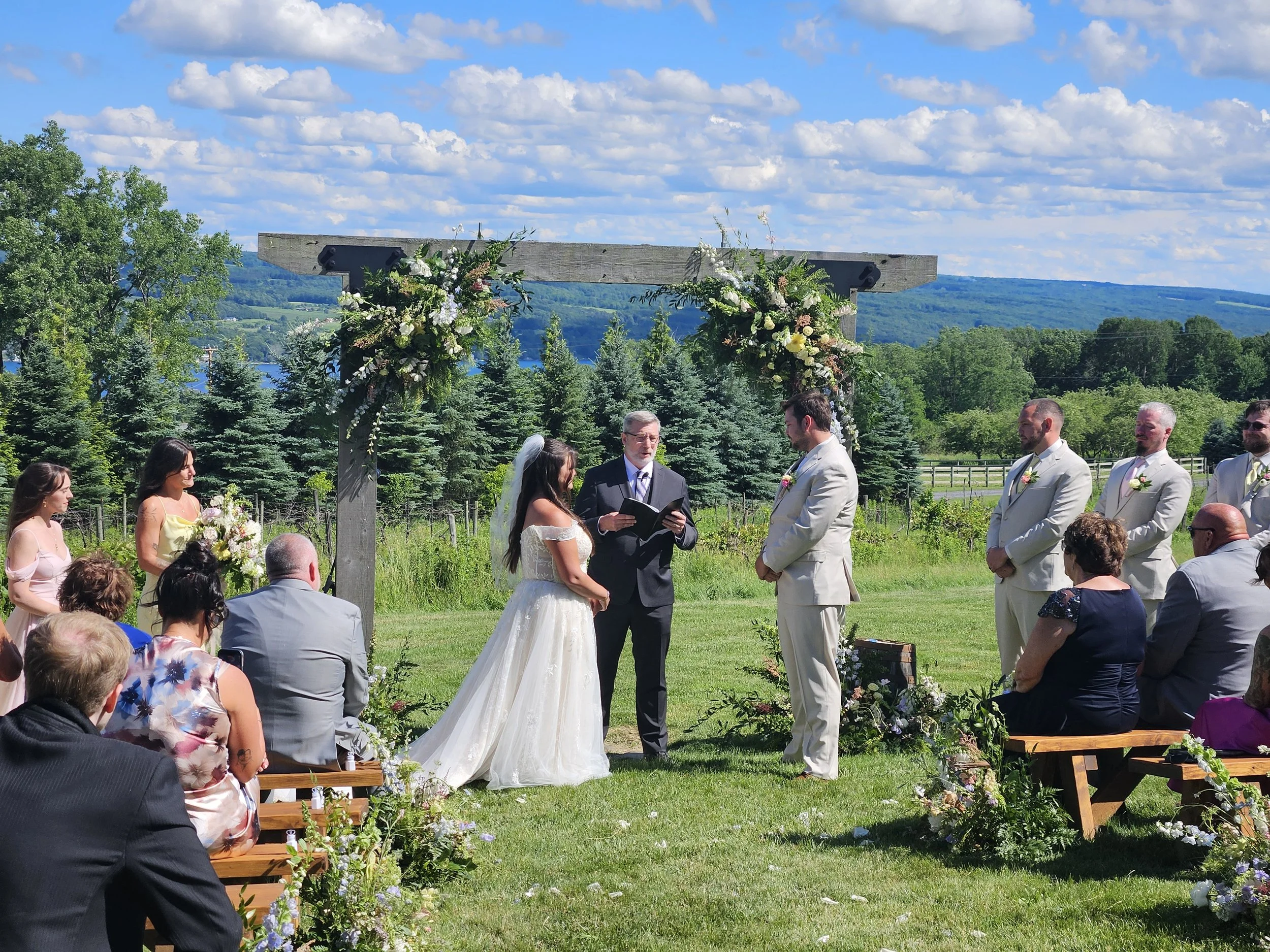 A wedding ceremony taking place outdoors on a grassy field under a clear blue sky with scattered clouds. The bride and groom are facing each other, with the officiant standing behind them. The ceremony is decorated with floral arrangements on a wooden arch and along the aisle, surrounded by guests seated on benches. In the background, there are lush green trees and a river with distant hills.