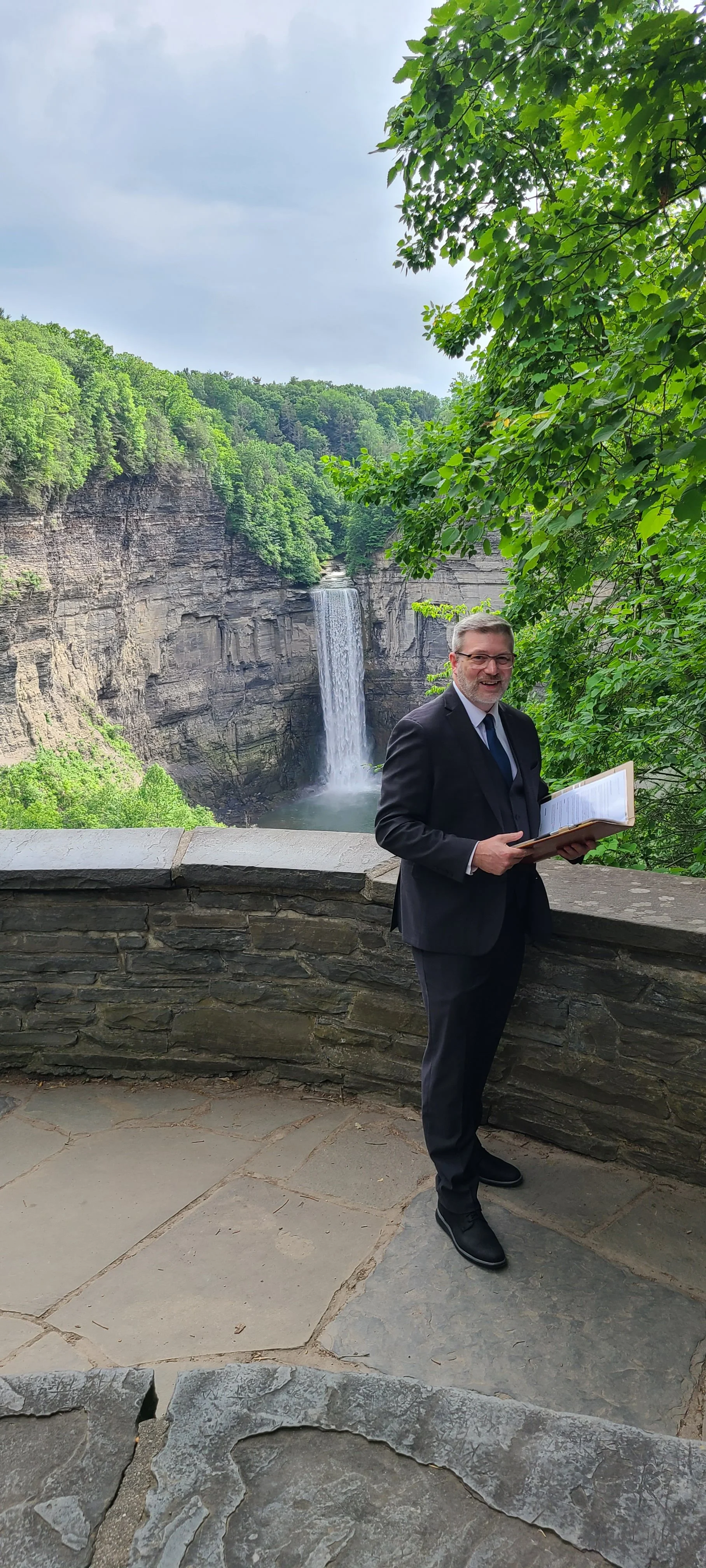 A man in a black suit holding a book or notebook, standing on a stone balcony with a waterfall and lush green trees in the background.