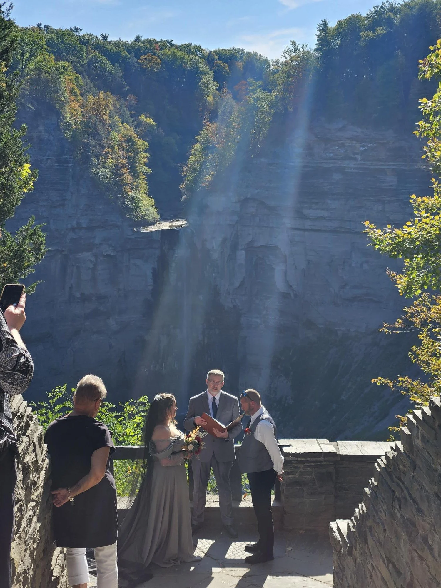 A wedding ceremony taking place outdoors at a scenic location surrounded by trees and cliffs, with a bride and groom exchanging vows and a few guests watching.