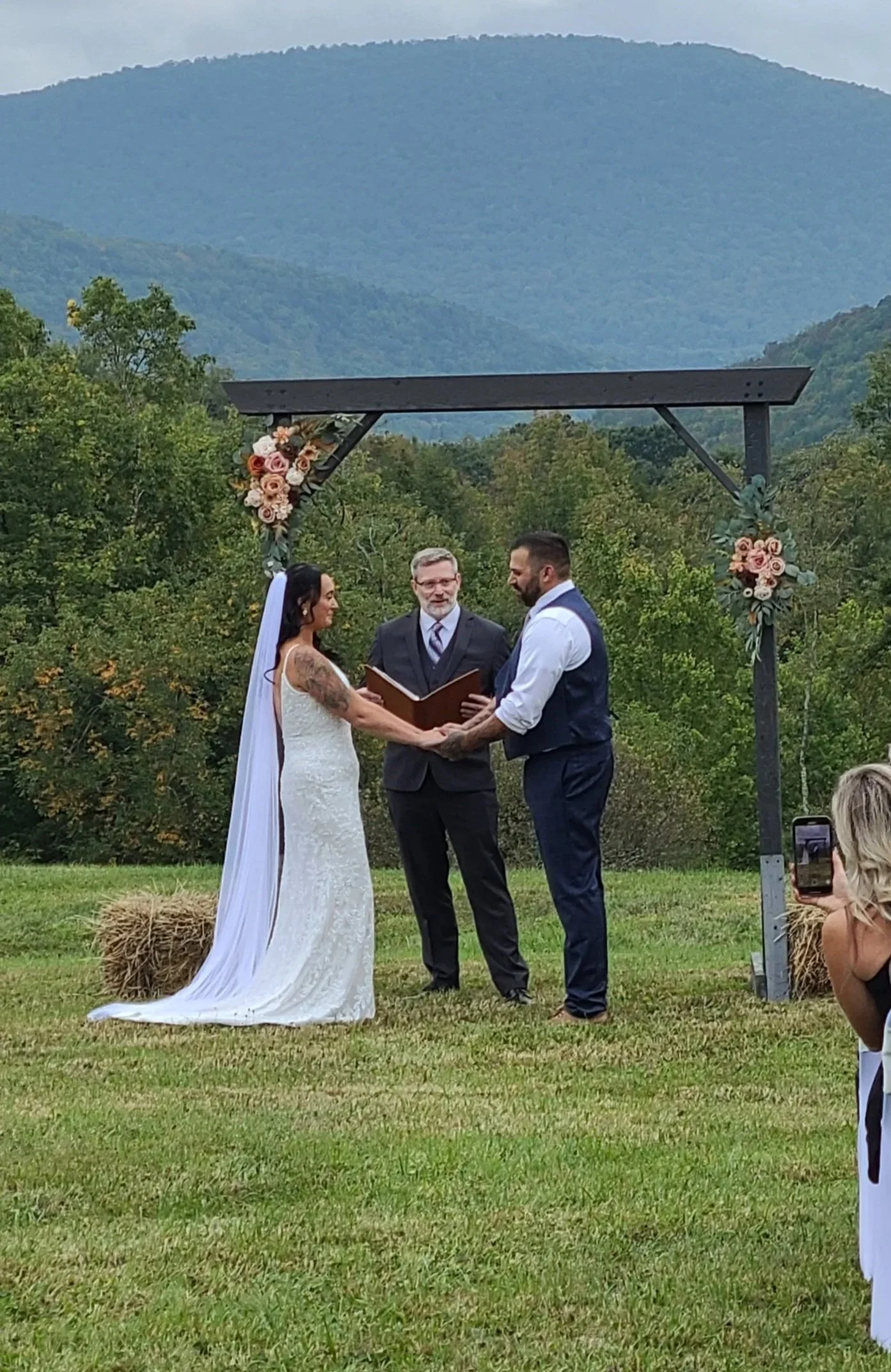 A wedding ceremony outdoors with a bride and groom holding hands, an officiant, and a floral arch against a backdrop of green trees and distant mountains.