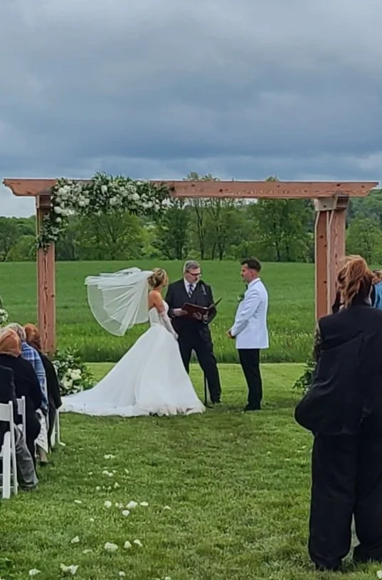 A wedding ceremony taking place outdoors under a wooden arbor decorated with flowers, with the bride and groom exchanging vows and guests seated on the lawn watching.