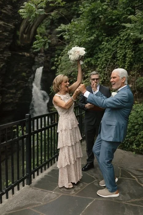A woman in a beige dress holding a bouquet of flowers overhead, celebrating with a man in a blue suit on a scenic outdoor pathway with a waterfall and lush green trees in the background.