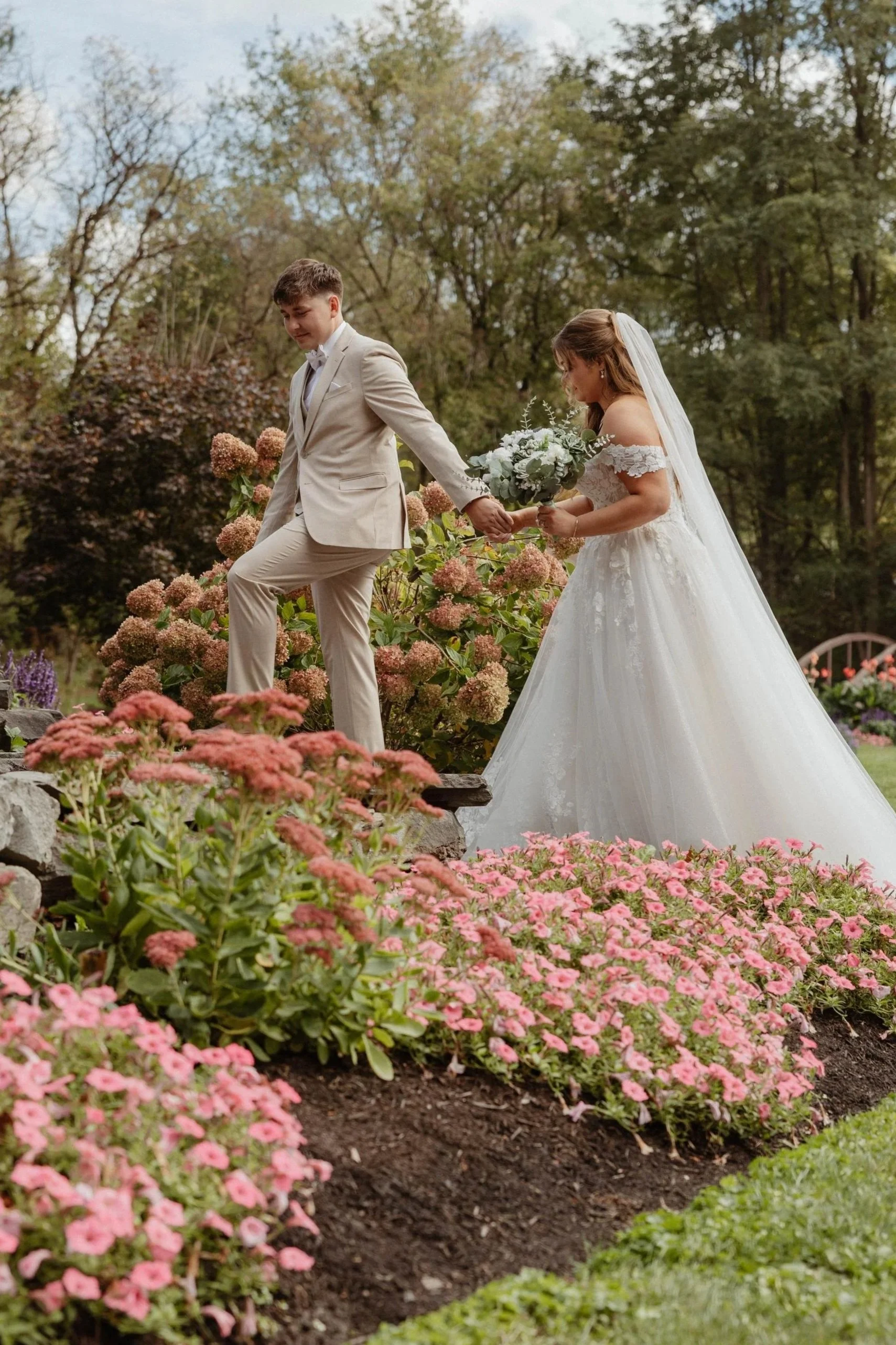A bride and groom holding hands outdoors among pink and green flowers, trees in the background, during a wedding.
