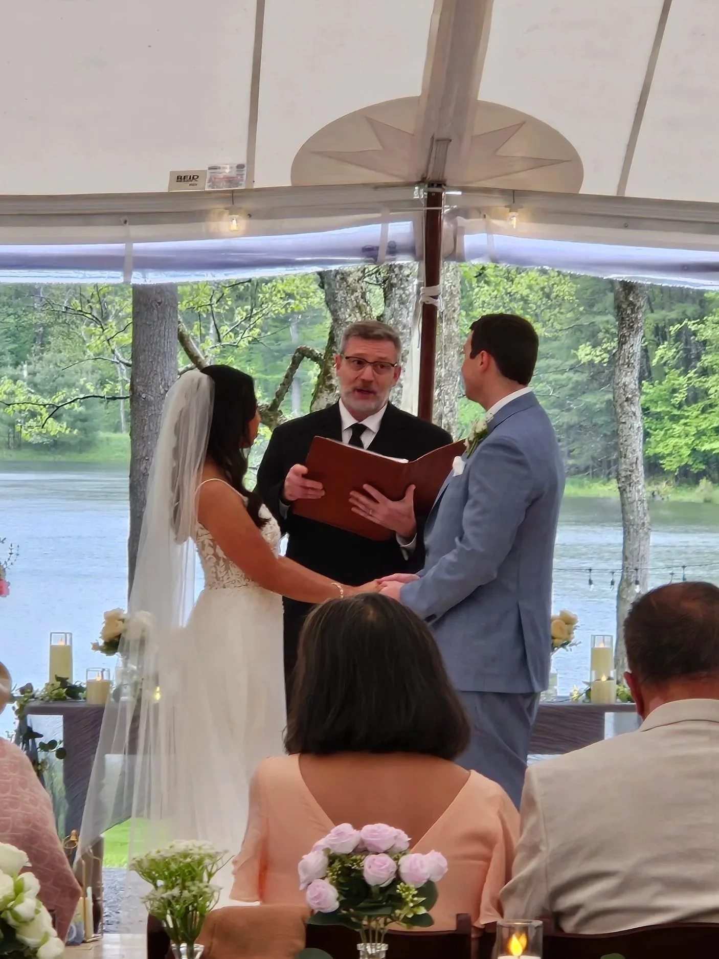 A bride and groom holding hands and facing each other during a wedding ceremony. The officiant, a man in a black suit, is holding a book and standing between them. The ceremony is outdoors near a lake, under a canopy decorated with candles and flowers, with trees and water visible in the background.