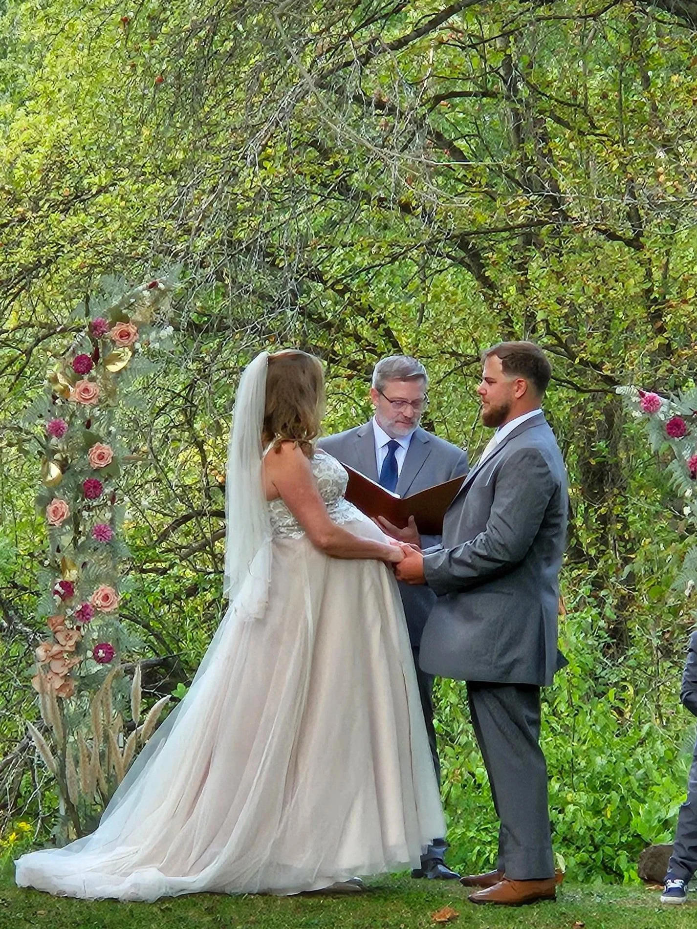 A couple getting married outdoors with a man officiating, surrounded by green trees and flowers.